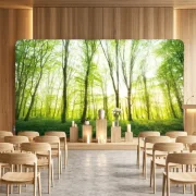 Eine moderne, holzgetäfelte Trauerhalle mit hellen Holzstühlen und einer großen, 400x230 cm großen Zipper-Wall im Stirnbereich, die ein helles Frühlingswald-Motiv hinter einer Urne zeigt.