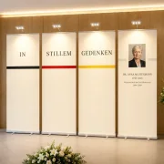 Ein vierteiliges elfenbeinfarbenes Roll-Up-Gedenkset wird in einem modernen Abschiedsraum von vier aufsteckbaren LED-Leuchten beleuchtet. Jedes Roll-Up besitzt eine eigene silberne Leuchte mit drei LED-Spots. Das warmweiße Licht hebt die Beschriftung „In stillem Gedenken“, die dezenten Streifen in Schwarz, Rot und Gold sowie die personalisierte Gedenkstele mit Porträt und Lebensdaten wirkungsvoll hervor.