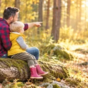 Vater und Sohn sitzen auf einem Baumstamm; der Vater zeigt auf einen hellen Weg durch den Wald.