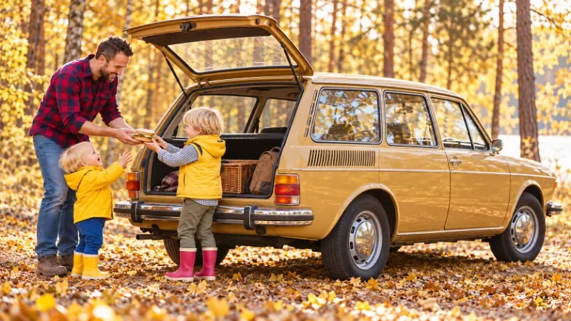 Ein Vater hilft seinem Sohn mit roten Gummistiefeln beim Einsteigen in einen historischen VW 412 Variant; der jüngere Bruder steht neben ihnen.