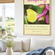 Hyperrealistische Wohnraumszene eines modernen friesischen Wohnzimmers mit heller, skandinavisch inspirierter Einrichtung und norddeutscher Wohnatmosphäre. Das christliche DIN-A2-Hochformat-Poster „Schöpfung“ mit einem Zitronenfalter auf einer rosafarbenen Blüte und dem inspirierenden Zitat von Jonathan Edwards hängt in einem hochwertigen Rahmen aus hellem Eichenholz über einem hellen Sofa. Natürliche Materialien, sanfte Farben und viel Tageslicht schaffen eine ruhige, einladende Atmosphäre und zeigen, wie stilvoll christliche Wanddekoration moderne Wohnräume bereichern kann.