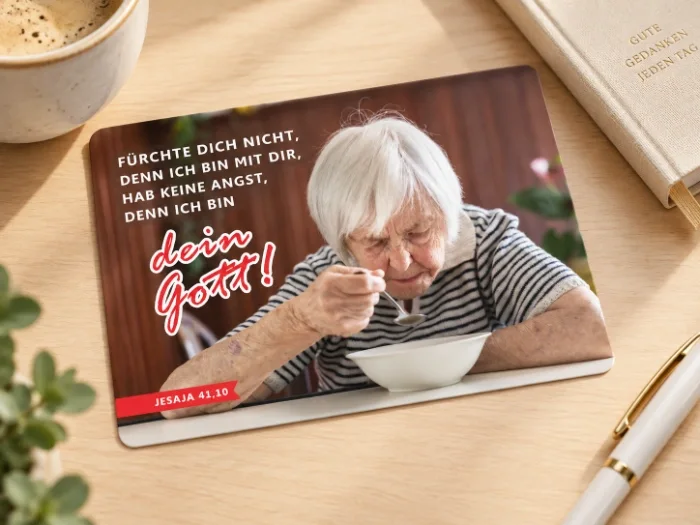 Produktfoto der Postkarte Jahreslosung 2027 mit älterer Frau allein am Tisch auf einem stilvollen Schreibtisch