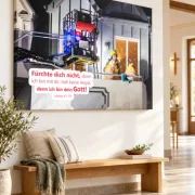 Großes Leinwandbild zur Jahreslosung 2027 mit einem Feuerwehrmann bei der Hilfe für ein Ehepaar, über einer Holzbank im Kirchenfoyer.
