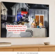 Leinwandbild Jahreslosung 2027 mit Feuerwehrmann und älterem Ehepaar in 80 × 60 cm über einem Sideboard mit Holzkreuz und Büchern.