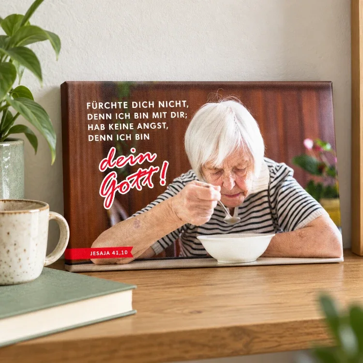 Leinwandbild Jahreslosung 2027 – Du bist nicht allein im Geschenkformat Kleines Leinwandbild zur Jahreslosung 2027 mit Seniorin beim Essen, auf einem Holzregal neben einer Tasse und einem Buch.