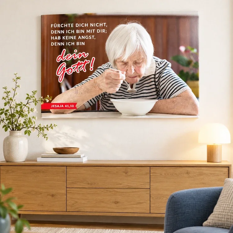 Jahreslosung 2027 auf Leinwand – Seniorinnen-Motiv im Wohnzimmer - 120 x 80 cm Leinwandbild Jahreslosung 2027 mit Seniorin und Bibelvers über einer Holzkommode, daneben eine Vase und eine Tischlampe.