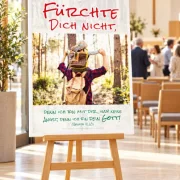 DIN-A1-Poster zur Jahreslosung 2027 auf einer Holzstaffelei in einem modernen Gemeindesaal