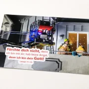 Freigestelltes DIN-A2-Poster zur Jahreslosung 2027 mit Feuerwehrmann und älterem Ehepaar