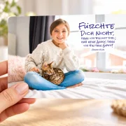 Hand hält eine Postkarte im DIN-A6-Format mit Mädchen, Katze und der Jahreslosung 2027