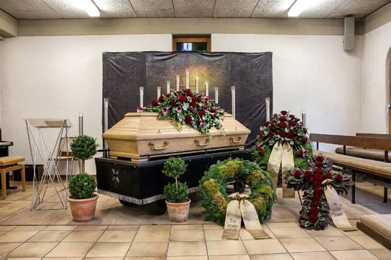 Würdevoll gestaltete Trauerhalle mit einem hellen Holzsarg auf einer Aufbahrung, geschmückt mit roten Rosen, Trauerkränzen und Kerzenleuchtern für Bestatter.