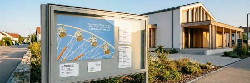 Ein moderner, großer Schaukasten aus Metall vor dem Holz- und Betongebäude einer Freien Evangelischen Gemeinde (FeG) am späten Nachmittag. Im Schaukasten hängt ein blaues Poster mit einem Riesenrad und Bibelvers sowie Infomaterialien. Keine Personen sichtbar.